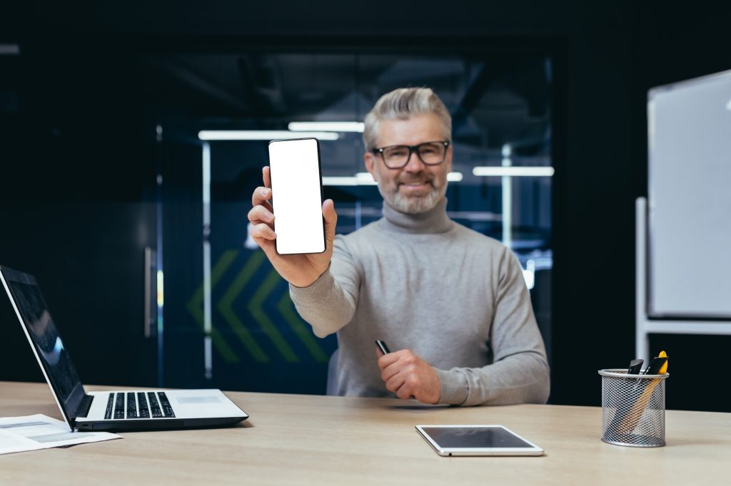 Successful businessman inside office showing phone with white screen free space to camera, mature